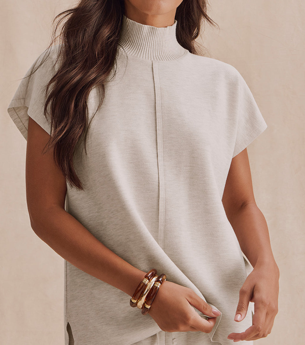 Woman wearing beige turtleneck sweater with gold and burgundy bangle bracelets against neutral background